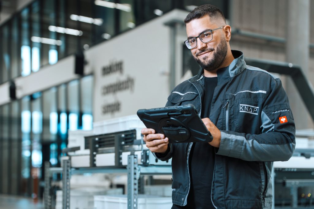 KNAPP customer service technician working inside a warehouse, smiling while performing system checks and providing hands‑on support for automated equipment.